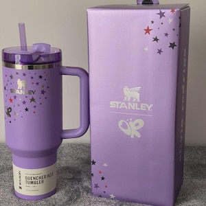 May include: A purple Stanley Quencher H2.0 tumbler with a handle and straw, next to its matching box. The tumbler and box are decorated with colorful star patterns. The tumbler has the Stanley logo and product information printed on it.