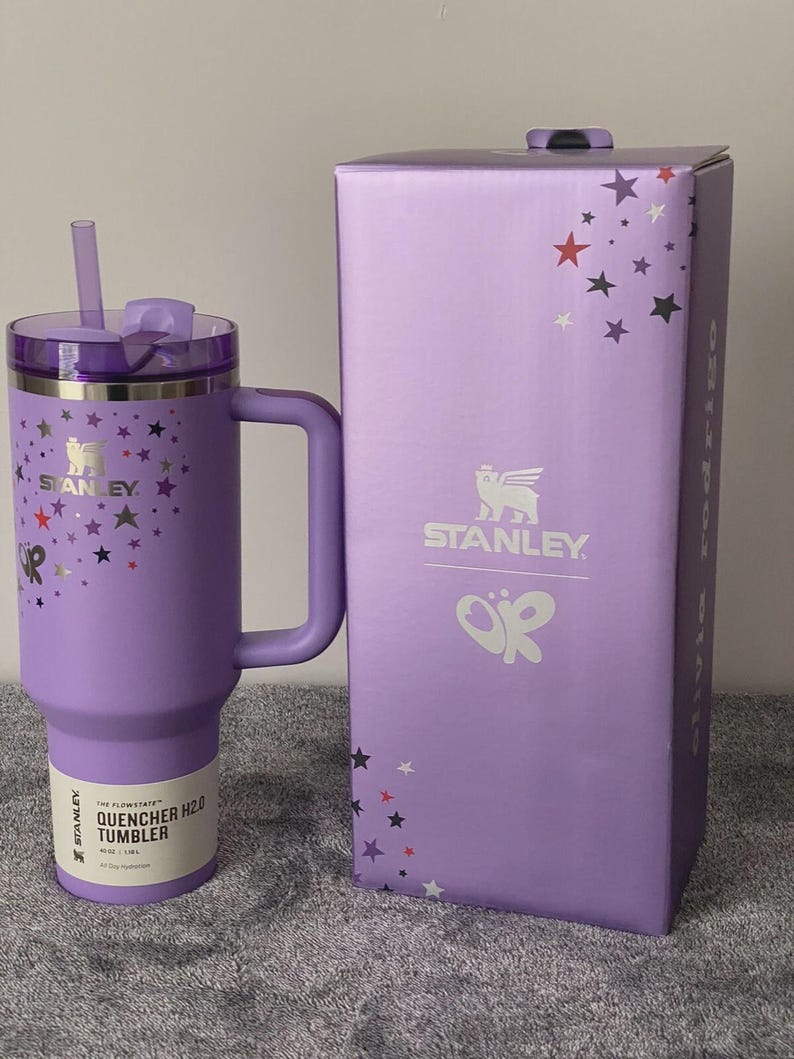 May include: A purple Stanley Quencher H2.0 tumbler with a handle and straw, next to its matching box. The tumbler and box are decorated with colorful star patterns. The tumbler has the Stanley logo and product information printed on it.