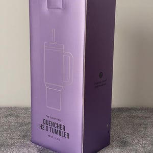 May include: A purple box featuring a line drawing of a tumbler with a handle and straw. The text on the box reads "THE FLOWSTATE QUENCHER H2.0 TUMBLER" and "40 OZ | 1.18 L". The box is on a gray surface.