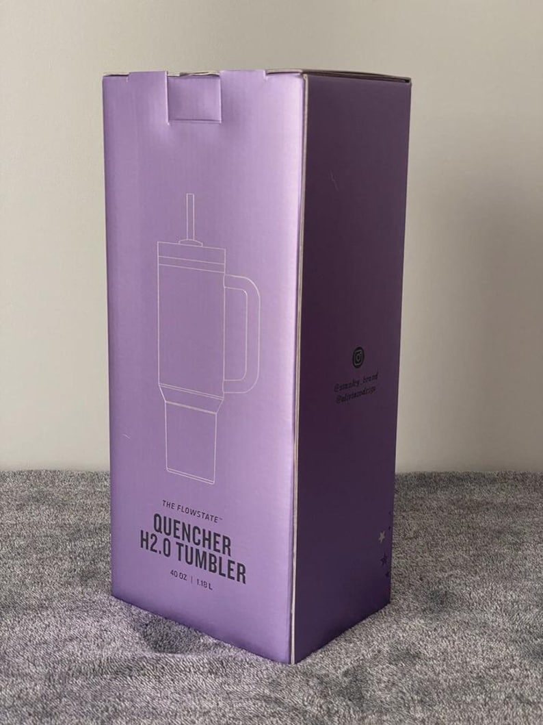 May include: A purple box featuring a line drawing of a tumbler with a handle and straw. The text on the box reads "THE FLOWSTATE QUENCHER H2.0 TUMBLER" and "40 OZ | 1.18 L". The box is on a gray surface.