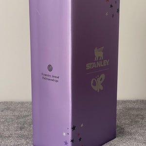 May include: A tall, rectangular box in a light purple hue. The box features the "STANLEY" logo with a Pegasus-like emblem and the initials "OR" in a butterfly design. The box also has star accents and social media handles.
