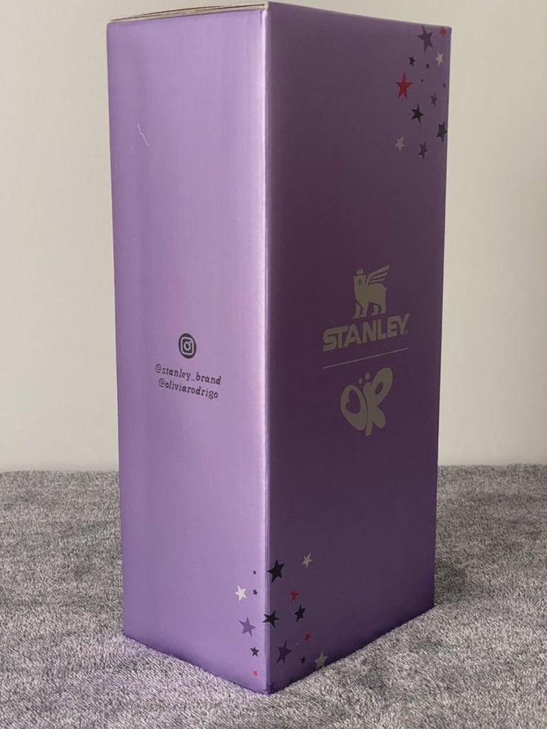 May include: A tall, rectangular box in a light purple hue. The box features the "STANLEY" logo with a Pegasus-like emblem and the initials "OR" in a butterfly design. The box also has star accents and social media handles.