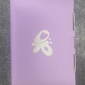 May include: A rectangular, lavender-colored box with a white, stylized butterfly logo. The box has a hinged side and a folded closure. The logo features a heart shape within the design. The box is on a gray surface.