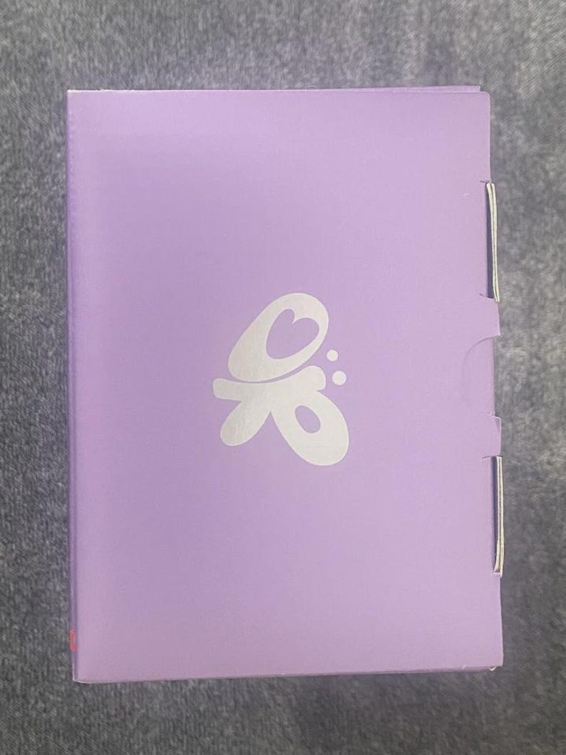 May include: A rectangular, lavender-colored box with a white, stylized butterfly logo. The box has a hinged side and a folded closure. The logo features a heart shape within the design. The box is on a gray surface.