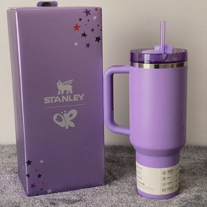 May include: A lavender Stanley tumbler with a handle and straw, next to its matching box. The box features the Stanley logo and star graphics. The tumbler has a capacity of 40 ounces.