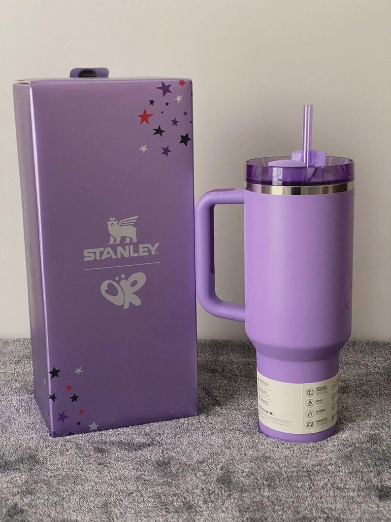 May include: A lavender Stanley tumbler with a handle and straw, next to its matching box. The box features the Stanley logo and star graphics. The tumbler has a capacity of 40 ounces.