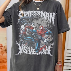 May include: Dark grey t-shirt with a graphic of Spider-Man and Venom in combat. The word "SPIDERMAN" is printed above the characters in a white, gothic font, with "VS VENOM" below. The shirt has a vintage, worn appearance.