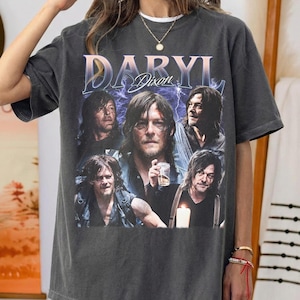 Puede incluir: Camiseta gris oscuro con un diseño gráfico de estilo vintage que presenta el nombre "DARYL DIXON" y un collage de imágenes de un hombre con pelo largo. El diseño incluye rayos en el fondo.