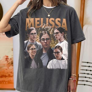 May include: Charcoal grey t-shirt with a graphic of a person wearing glasses, and the name "MELISSA King" in a gold font. The graphic includes multiple images of the person. The shirt has a vintage, worn appearance.