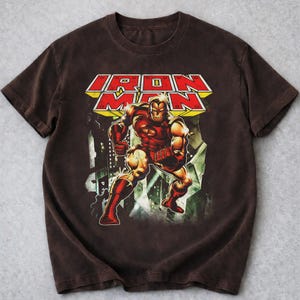 May include: Brown t-shirt with a graphic of Iron Man in a dynamic pose. The design includes the words "IRON MAN" in a bold, red and yellow font. The artwork depicts Iron Man in his classic red and gold armour.