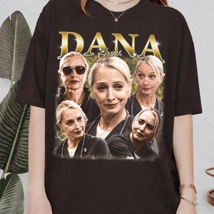Op de afbeelding: Donkerbruin T-shirt met de naam "DANA Evans" in gouden letters. Het T-shirt heeft een collage van afbeeldingen van een vrouw met een zonnebril en een bril. De hals is rond.