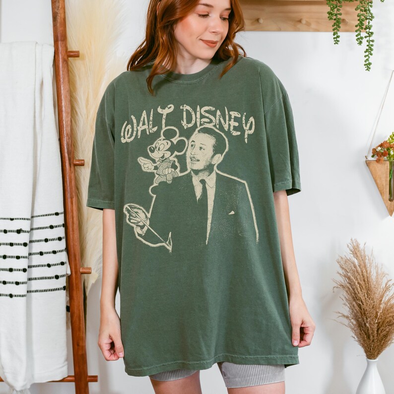 May include: An olive green short-sleeve t-shirt featuring a distressed cream-colored graphic print of Walt Disney and Mickey Mouse. The text "WALT DISNEY" is printed in a stylized font above the figures, with the design centered on the chest.