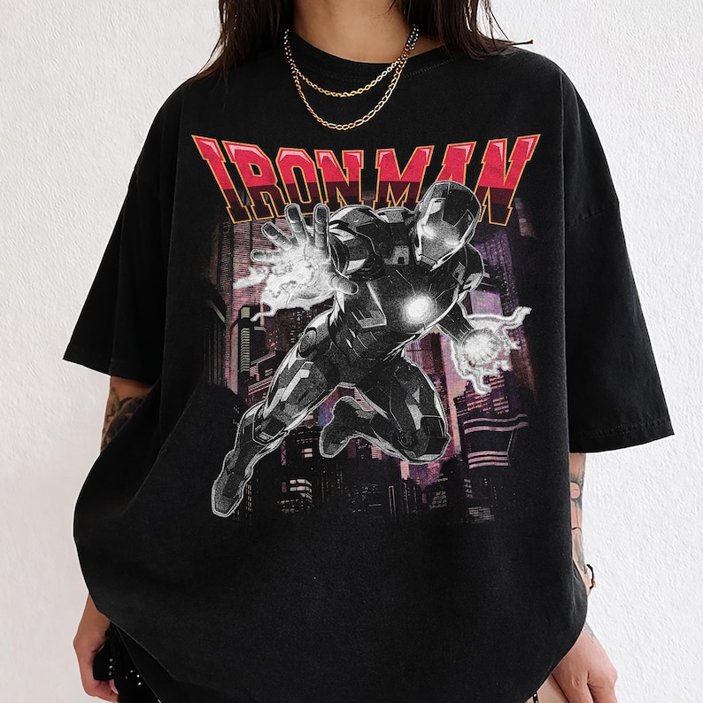 May include: Black t-shirt featuring a graphic of Iron Man in action, set against a cityscape backdrop. The text "IRON MAN" is displayed in red and gold. The shirt has a vintage, worn look.