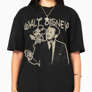 May include: A black short-sleeve t-shirt featuring a graphic print of Walt Disney and Mickey Mouse. The text "WALT DISNEY" is printed in a distressed, light-colored font above the figures. The design includes a portrait of Walt Disney.