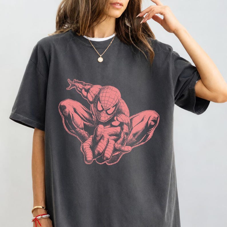 May include: A charcoal grey short-sleeve t-shirt featuring a large, centered graphic print of the comic book character Spider-Man in a crouching pose. The graphic is rendered in a muted, dusty pink color with detailed black line work.