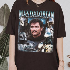 K&ouml;nnte beinhalten: Dunkelbraunes T-Shirt mit einer Grafik des Mandalorianers, mit Bildern der Figur und Baby Yoda. Das Wort "MANDALORIAN" ist oben in einer blauen, stilisierten Schriftart aufgedruckt.