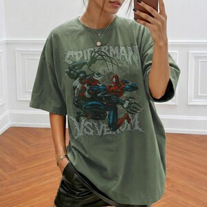 May include: An olive green oversized t-shirt with a graphic of Spider-Man and Venom in combat. The text "SPIDERMAN" is printed above the characters. The shirt has short sleeves and a loose fit.