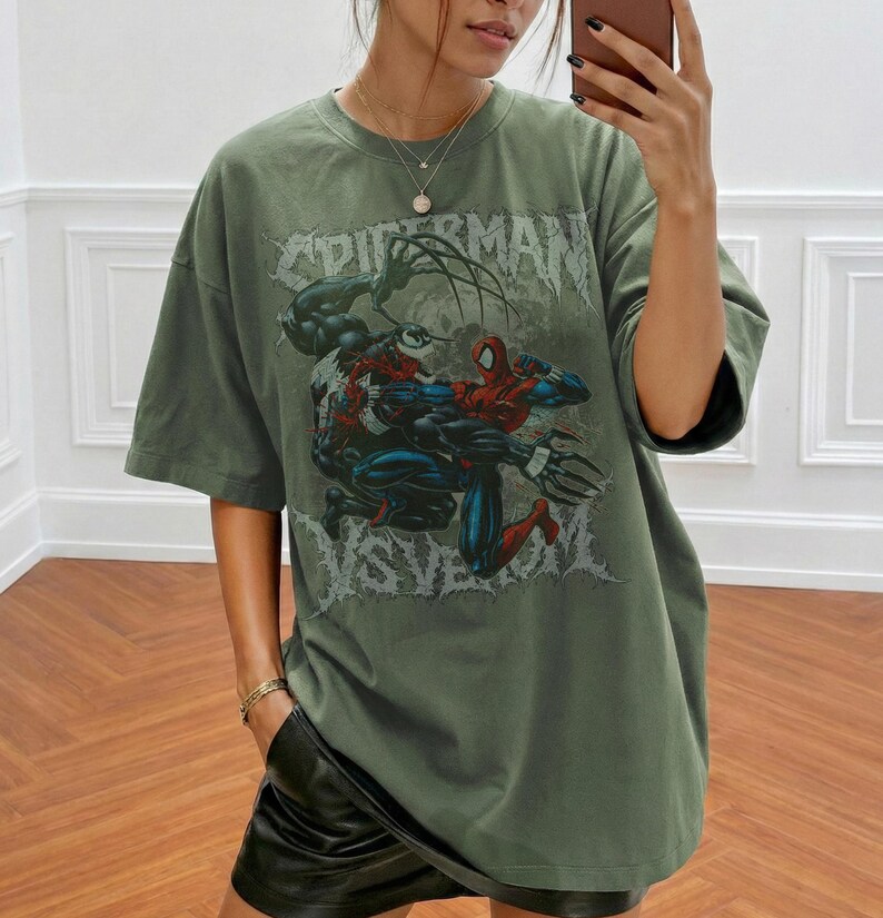 May include: An olive green oversized t-shirt with a graphic of Spider-Man and Venom in combat. The text "SPIDERMAN" is printed above the characters. The shirt has short sleeves and a loose fit.