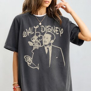 May include: A charcoal gray short-sleeve t-shirt featuring a distressed cream-colored graphic print of Walt Disney and Mickey Mouse. The text "WALT DISNEY" is printed in a stylized font above the figures, with Walt Disney holding a conductor baton.