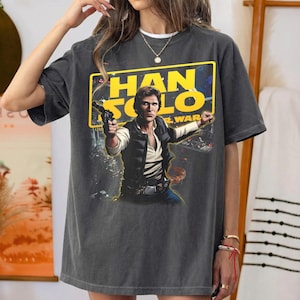 Pode incluir: Uma t-shirt cinzento-carvão de manga curta com uma impressão gráfica de Han Solo de Star Wars. O design inclui a personagem a segurar um blaster, sobre um fundo retangular amarelo com o texto HAN SOLO em letras amarelas a negrito.