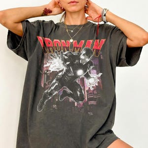 May include: Dark gray t-shirt featuring the text "IRONMAN" in red and a graphic of Iron Man. The image shows Iron Man in a dynamic pose with energy effects, set against a cityscape background.