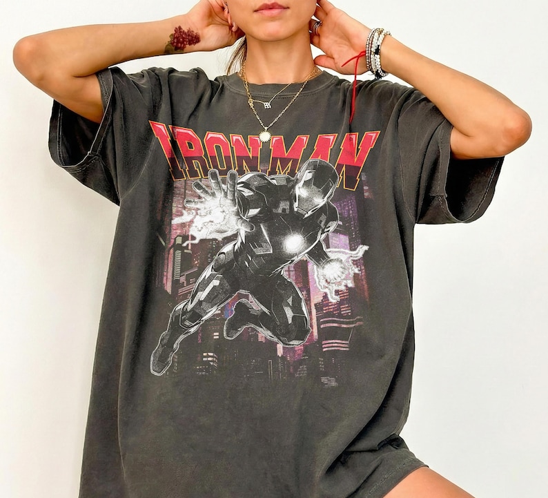 May include: Dark gray t-shirt featuring the text "IRONMAN" in red and a graphic of Iron Man. The image shows Iron Man in a dynamic pose with energy effects, set against a cityscape background.