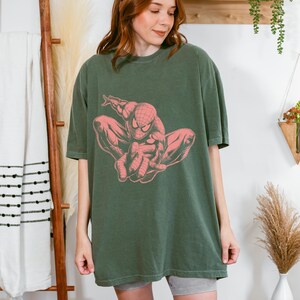May include: An olive green short-sleeved t-shirt featuring a large, centered graphic print of the comic book character Spider-Man in a crouching pose. The graphic is rendered in a muted coral pink color, contrasting against the dark green fabric.
