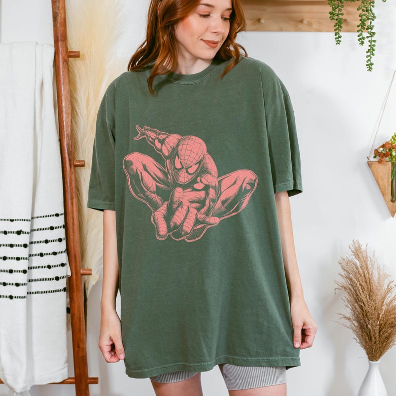 May include: An olive green short-sleeved t-shirt featuring a large, centered graphic print of the comic book character Spider-Man in a crouching pose. The graphic is rendered in a muted coral pink color, contrasting against the dark green fabric.