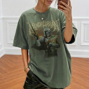 K&ouml;nnte beinhalten: Olivgr&uuml;nes T-Shirt mit einer Grafik von The Mandalorian, der eine Waffe und Grogu h&auml;lt. Der Text "MANDALORIAN" wird in einer stilisierten Schriftart &uuml;ber den Figuren angezeigt. Das Shirt hat kurze &Auml;rmel und eine lockere Passform.