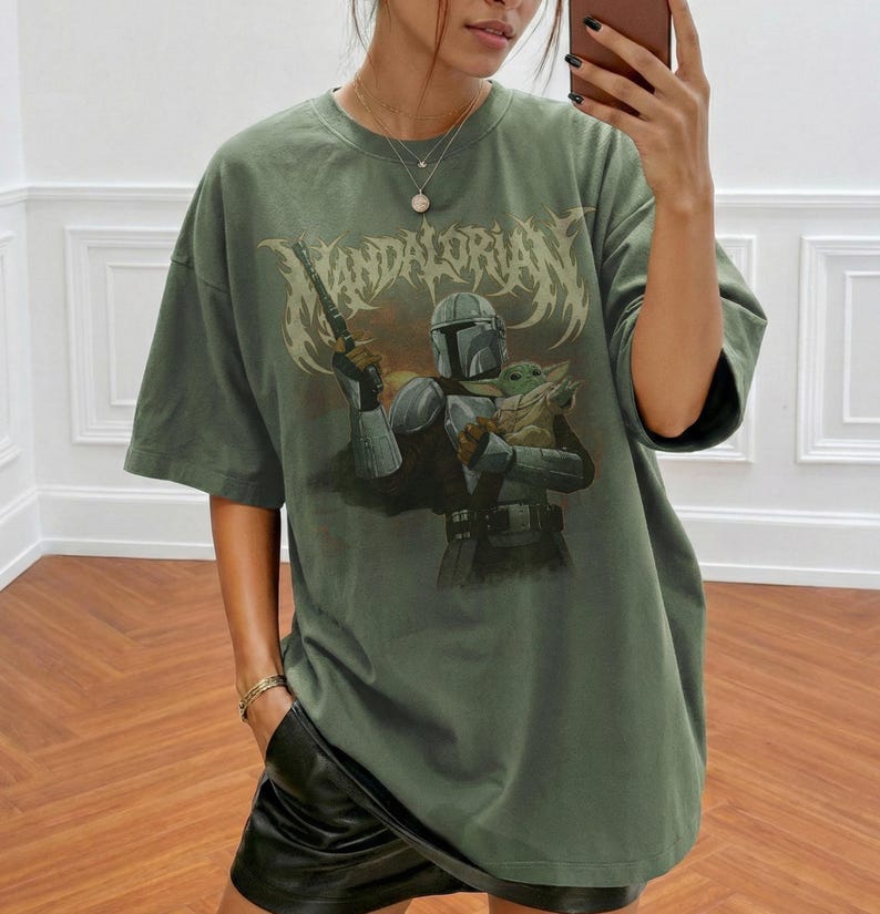 K&ouml;nnte beinhalten: Olivgr&uuml;nes T-Shirt mit einer Grafik von The Mandalorian, der eine Waffe und Grogu h&auml;lt. Der Text "MANDALORIAN" wird in einer stilisierten Schriftart &uuml;ber den Figuren angezeigt. Das Shirt hat kurze &Auml;rmel und eine lockere Passform.