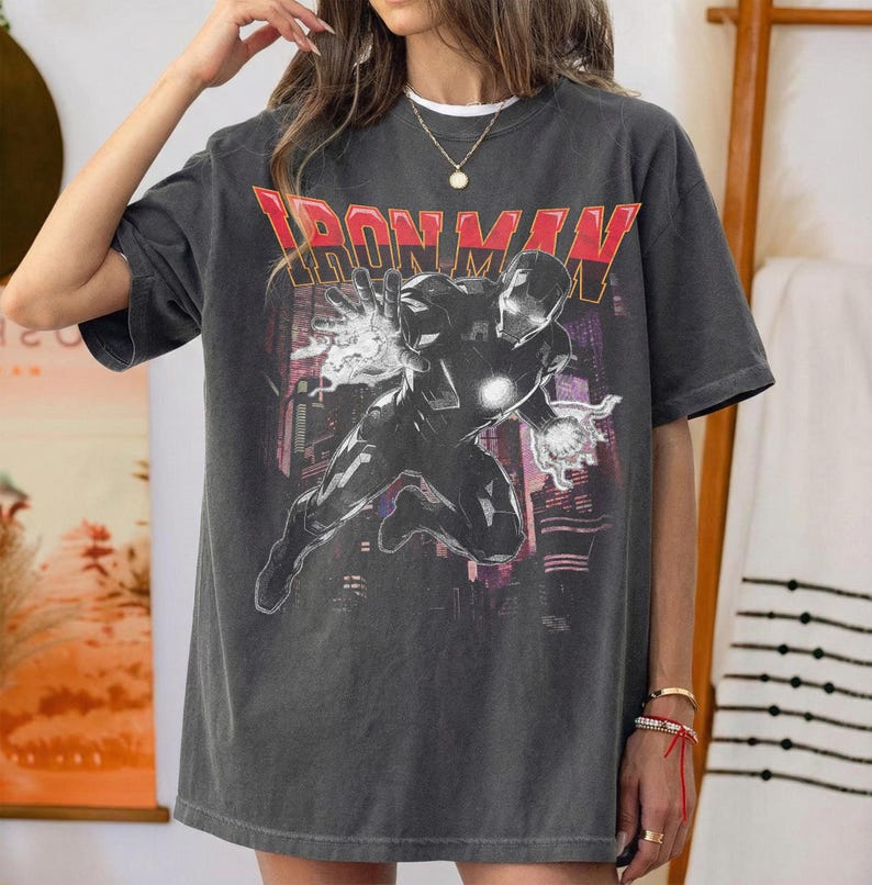 May include: Dark gray t-shirt featuring the text "IRON MAN" in red block letters above a graphic of Iron Man in action. The graphic shows Iron Man with his hands outstretched, with a cityscape in the background.
