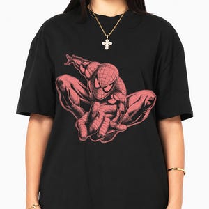 May include: A black short-sleeved t-shirt featuring a large, detailed graphic print of a crouching superhero character in a muted red tone. The character has a web-patterned mask and is depicted in a dynamic, forward-leaning pose on the chest.