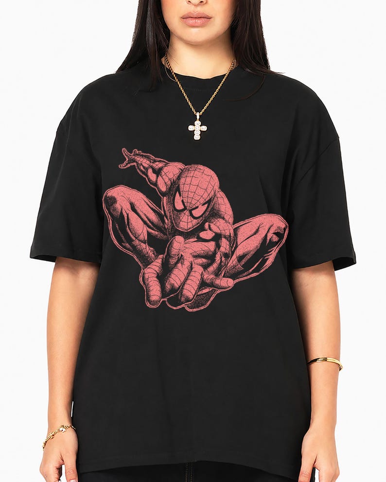 May include: A black short-sleeved t-shirt featuring a large, detailed graphic print of a crouching superhero character in a muted red tone. The character has a web-patterned mask and is depicted in a dynamic, forward-leaning pose on the chest.