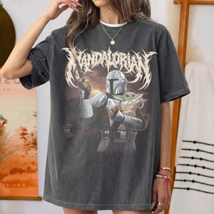 K&ouml;nnte beinhalten: Dunkelgraues T-Shirt mit einer Grafik von The Mandalorian, der Grogu h&auml;lt, mit dem Wort "Mandalorian" in einer stilisierten Schriftart. Das Design verwendet Grau-, Braun- und Wei&szlig;t&ouml;ne. Das T-Shirt hat kurze &Auml;rmel und eine lockere Passform.
