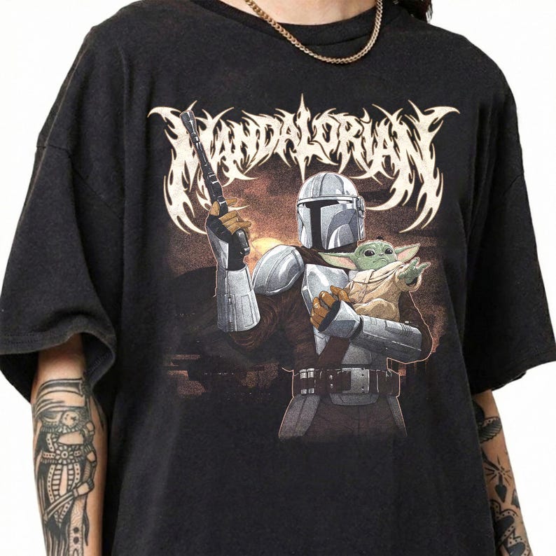 K&ouml;nnte beinhalten: Schwarzes T-Shirt mit einer Grafik von The Mandalorian, der eine Waffe h&auml;lt, und Grogu. Das Design enth&auml;lt das Wort "Mandalorian" in einer stilisierten Schriftart. Das Kunstwerk ist vor einem braunen und orangefarbenen Hintergrund platziert.