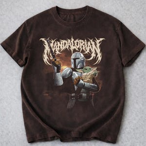 K&ouml;nnte beinhalten: Braunes T-Shirt mit einer Grafik eines Mandalorianer-Charakters, der eine Waffe und eine kleine gr&uuml;ne Kreatur h&auml;lt. Das Wort "MANDALORIAN" ist in einer stilisierten Schriftart &uuml;ber den Figuren aufgedruckt. Das Shirt hat kurze &Auml;rmel.
