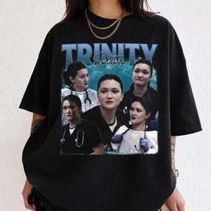 Könnte beinhalten: Schwarzes T-Shirt mit einem Collage-Bild einer Person, mit dem Namen "TRINITY Santos" in blauer Schrift. Die Person trägt medizinische Kleidung und ein Stethoskop. Das T-Shirt hat einen Rundhalsausschnitt.