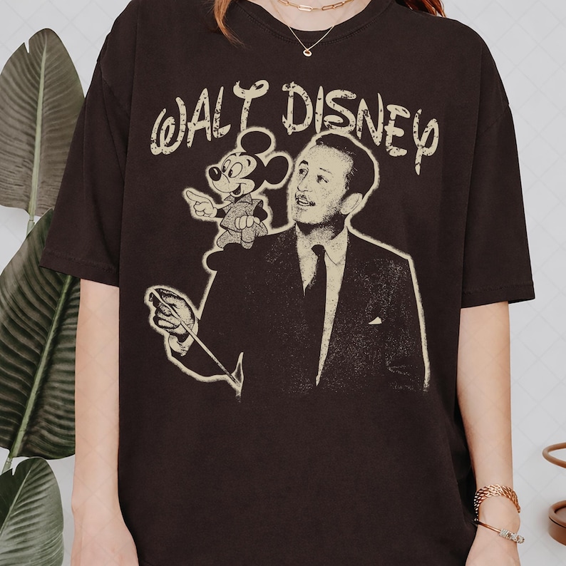May include: A dark brown short-sleeve t-shirt featuring a distressed cream-colored graphic print of Walt Disney and Mickey Mouse. The text WALT DISNEY is printed in a stylized font above the figures, with Walt Disney holding a conductor baton.