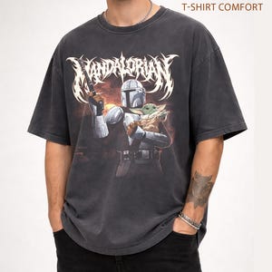 K&ouml;nnte beinhalten: Dunkelgraues T-Shirt mit einer Grafik von The Mandalorian mit einer Waffe und Grogu. Der Text "MANDALORIAN" befindet sich &uuml;ber dem Bild. Das Shirt ist mit "PEPPER T-SHIRT COMFORT" gekennzeichnet.