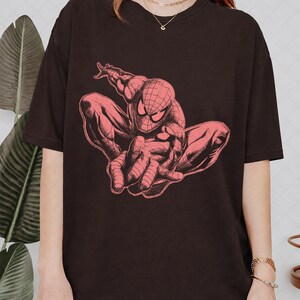 May include: A black short-sleeved t-shirt featuring a large, detailed graphic print of the character Spider-Man in a crouching pose. The illustration is rendered in a monochromatic reddish-pink tone, highlighting the character's web-patterned suit.