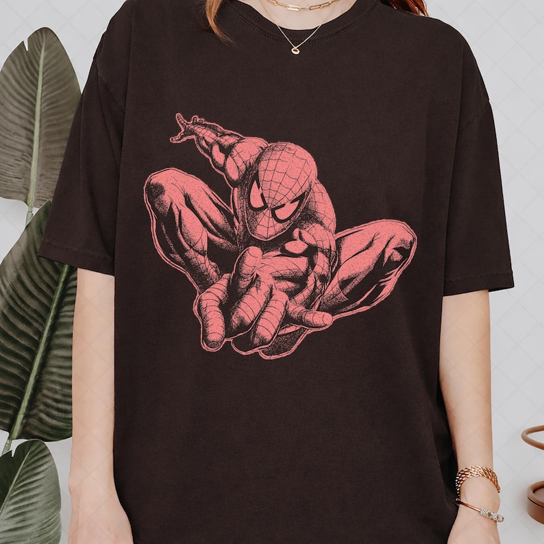 May include: A black short-sleeved t-shirt featuring a large, detailed graphic print of the character Spider-Man in a crouching pose. The illustration is rendered in a monochromatic reddish-pink tone, highlighting the character's web-patterned suit.