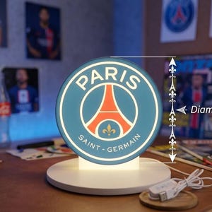 Peut inclure: Une lampe circulaire avec le logo du Paris Saint-Germain au design bleu et orange, dotée d'une base blanche et d'un câble d'alimentation USB. Un graphique indique un diamètre de 16 cm. L'emblème affiche la tour Eiffel et une fleur de lys.