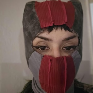 May include: A balaclava constructed from panels of dark gray, light gray, and deep red ribbed fabric. The headgear features a large cutout for the eyes and nose, with visible raw, frayed edges along the seams and the central opening of the mask.