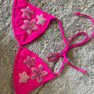 Può includere: Un top bikini rosa acceso con decorazioni floreali. Il top a triangolo presenta fiori ricamati su ogni coppa, con lacci per una vestibilità regolabile. Il bikini è realizzato in un tessuto liscio e tinta unita.