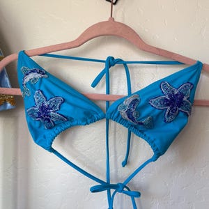 May include: A turquoise bikini top with beaded dolphin and flower embellishments. The triangle top is suspended from a pink hanger. The embellishments are in shades of blue and silver.