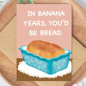May include: A greeting card with the text "IN BANANA YEARS, YOU'D BE BREAD." The card features an illustration of a loaf of bread in a turquoise baking dish. The card is on a wooden plate with a brown envelope.