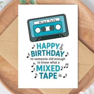 May include: A birthday card with a teal cassette tape illustration and the words "Happy Birthday" and "Mixed Tape." The card is on a wooden tray with a brown envelope.