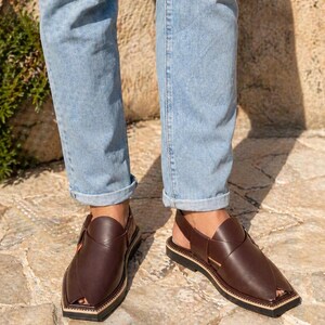 May include: Brown leather sandals with a distinctive design. The sandals feature an ankle strap and a square toe. The soles are black with a beige border. The sandals are worn with light blue jeans.