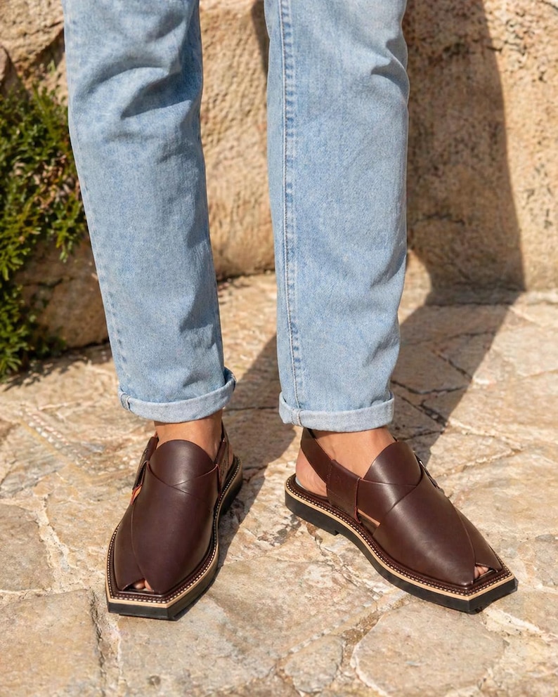 May include: Brown leather sandals with a distinctive design. The sandals feature an ankle strap and a square toe. The soles are black with a beige border. The sandals are worn with light blue jeans.
