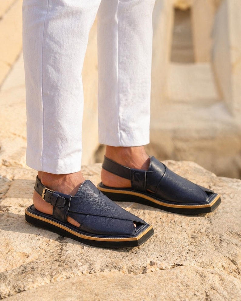 May include: Navy blue leather sandals with a square toe and an ankle strap. The sandals have a black sole with a tan trim and a gold buckle. The sandals are worn with white trousers.